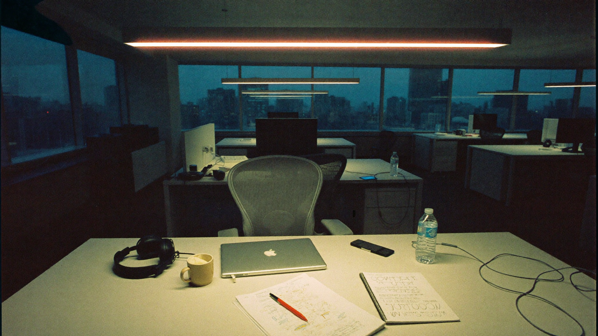 A single lit workstation in an otherwise dark open-plan office at night: closed laptop, headphones, an open notebook with hand-written notes, a cold cup of coffee. The empty chair pushed back from the desk.